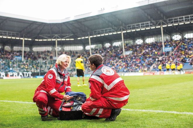 Foto: Zwei Mitarbeiter des DRK-Sanitätsdienstes tauschen sich am Spielfeldrand fachlich miteinander aus. Der Sanitätsrucksack liegt geöffnet zwischen den beiden.